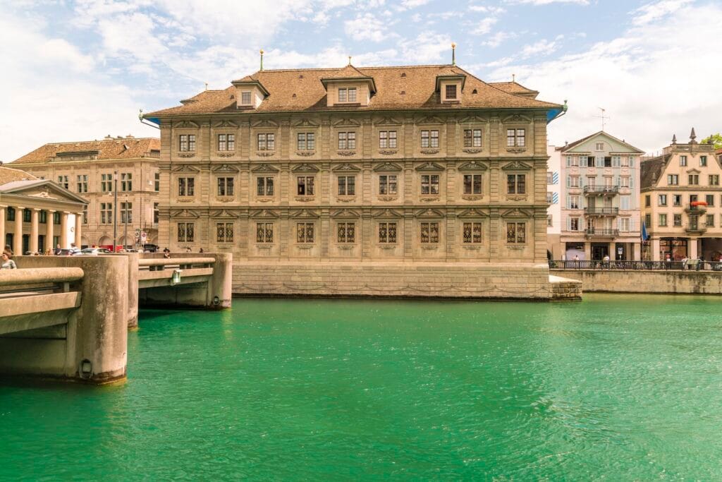 Здание швейцарского банка в центре города у реки — иллюстрация темы «Сколько стоит открыть счет в швейцарском банке», стоимость обслуживания, требования банков и финансовые условия в Швейцарии.